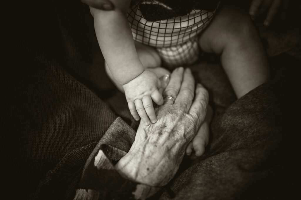 Mâna unui bebeluș sprijinită pe mâna ridată a unei bunici, fotografie alb-negru